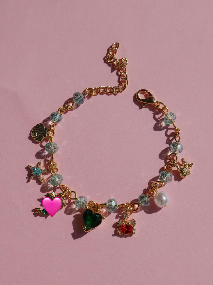 A gold-colored brass bracelet with multiple charms, including a heart-shaped pendant with a green and red design, set on a pink background.