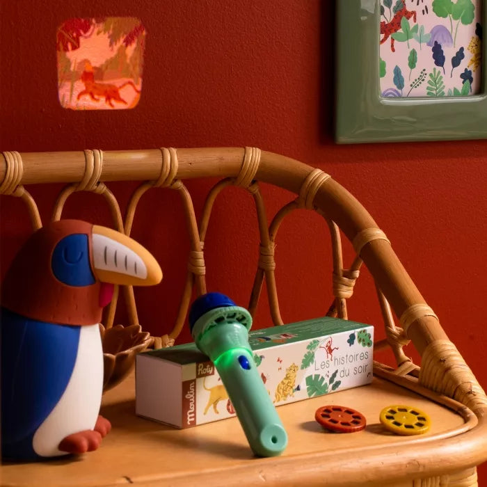 Toy toucan and green projector torch toy on a wooden surface with a red wall and wicker chair in the background.