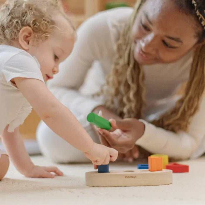 Child playing with wooden blocks on a mat, assisted by an adult.