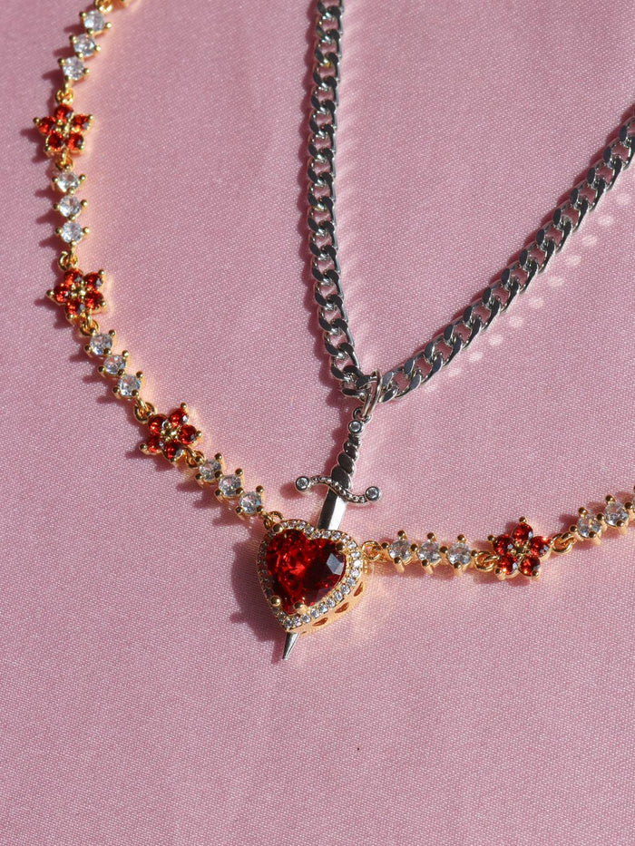 Two necklaces, one with a sword-shaped pendant and red stone accents, and the other a solid gold-tone chain, displayed on a pink background.